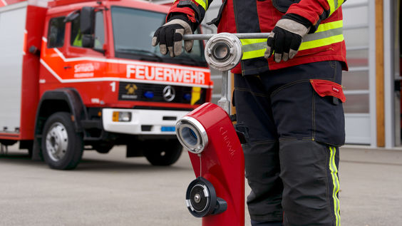 Bild von rotem Hydranten mit Feuerwehrmann, der vor einem Feuerwehrwagen steht und den Hydranten bedient und aufdreht.